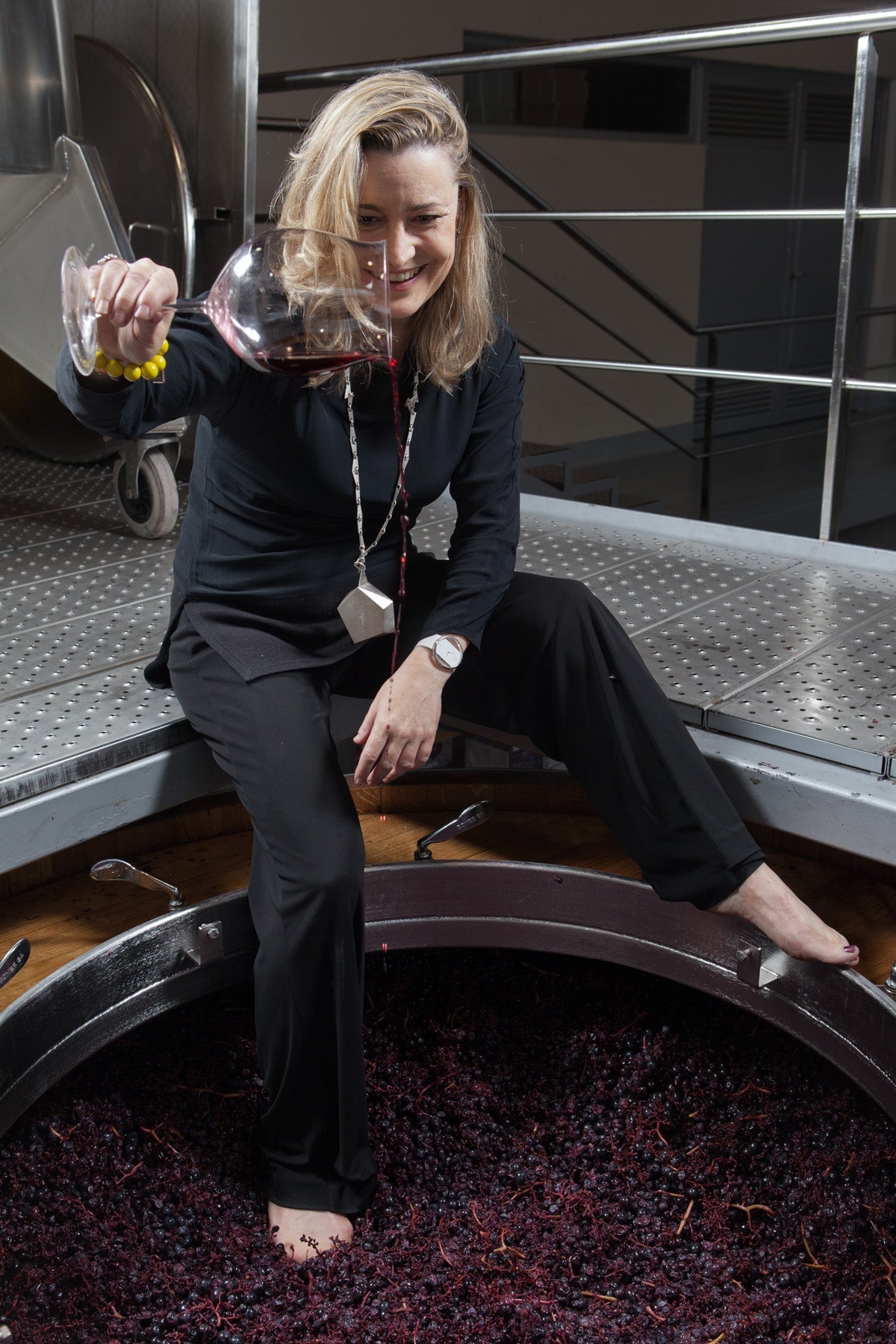 Winzerin sitzt barfuss am Rand eines offenen Gärbottichs voller Maische und schwenkt ein Glas Rotwein